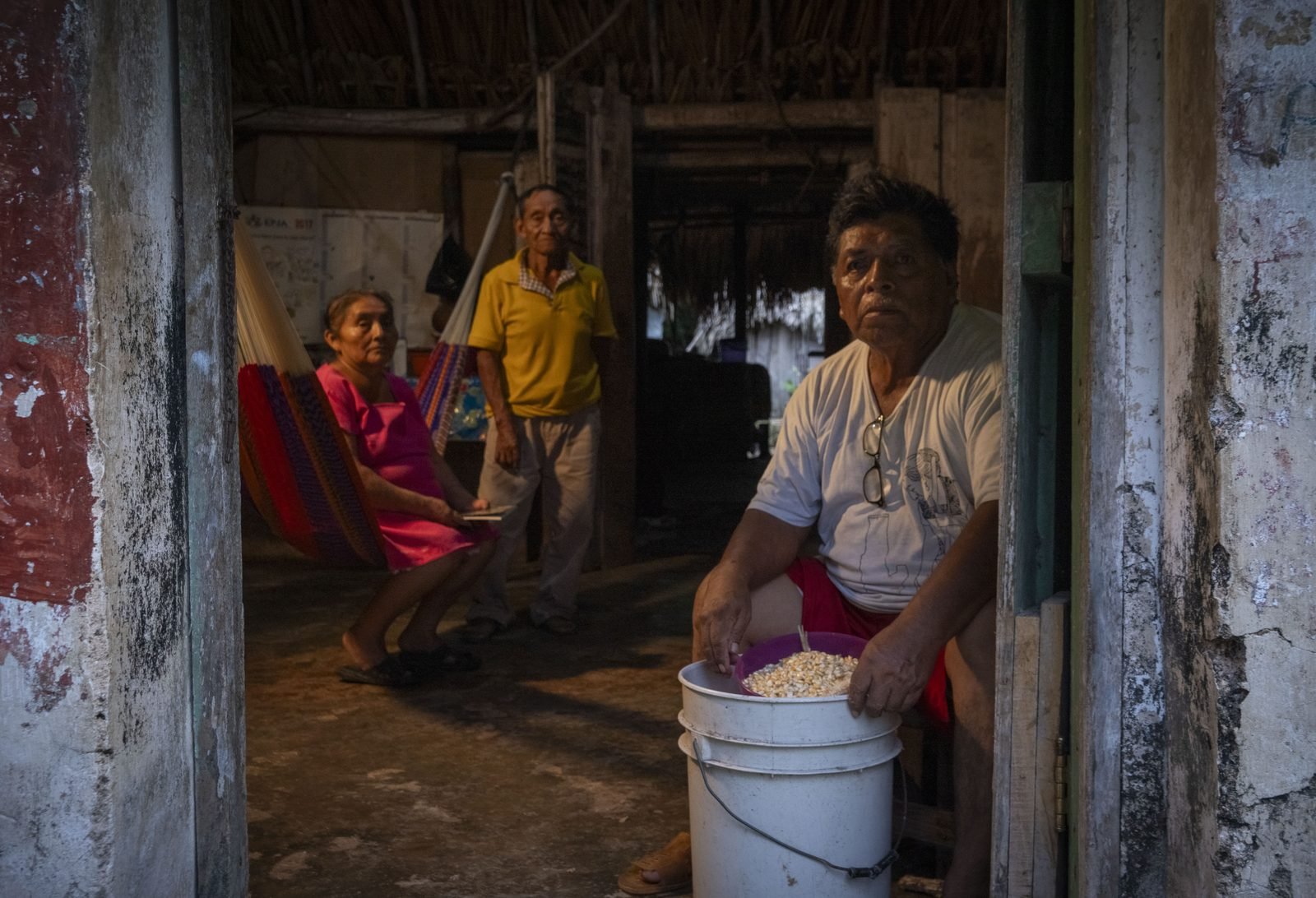 Familia maya en su hogar; al frente, un hombre con un balde de maíz de la milpa